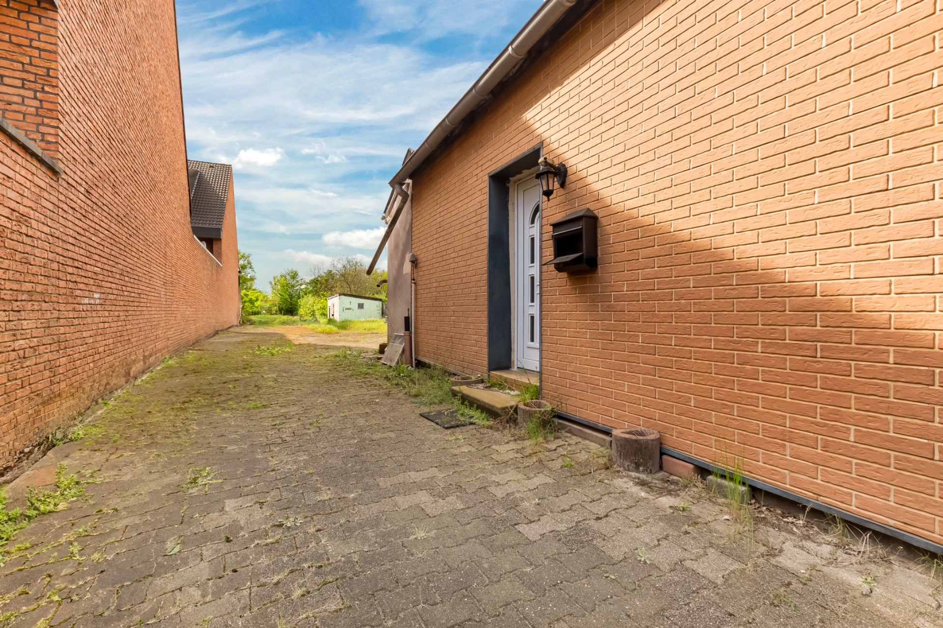 Gepflasterter Weg zwischen zwei Backsteingebäuden, mit Haustür und Briefkasten rechts.