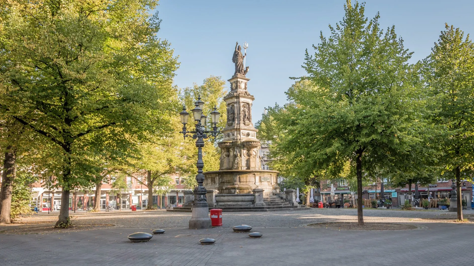 Ein steinerner Brunnen mit Statue steht auf einem von Bäumen umgebenen Platz.