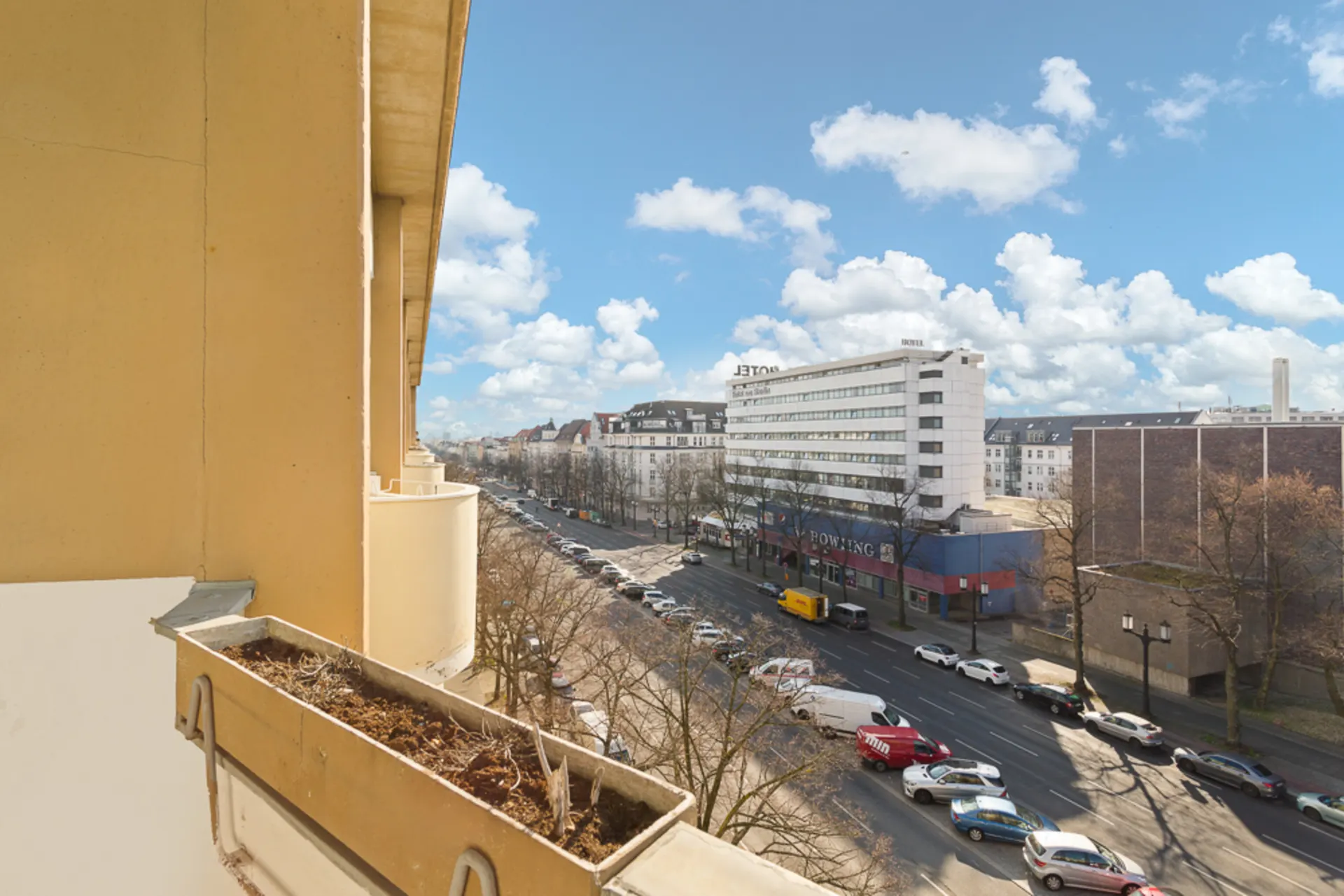 Blick von einem Balkon auf eine Straße mit Autos, Bäumen und Gebäuden bei blauem Himmel.