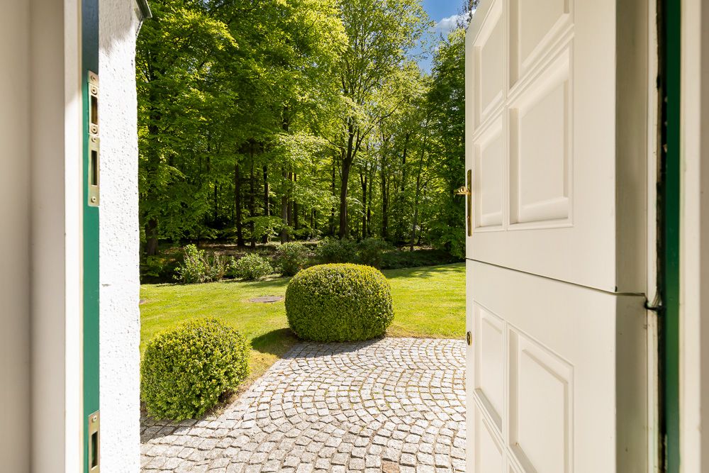 Blick durch eine offene Tür auf einen gepflasterten Weg und einen grünen Garten mit Büschen und Bäumen.