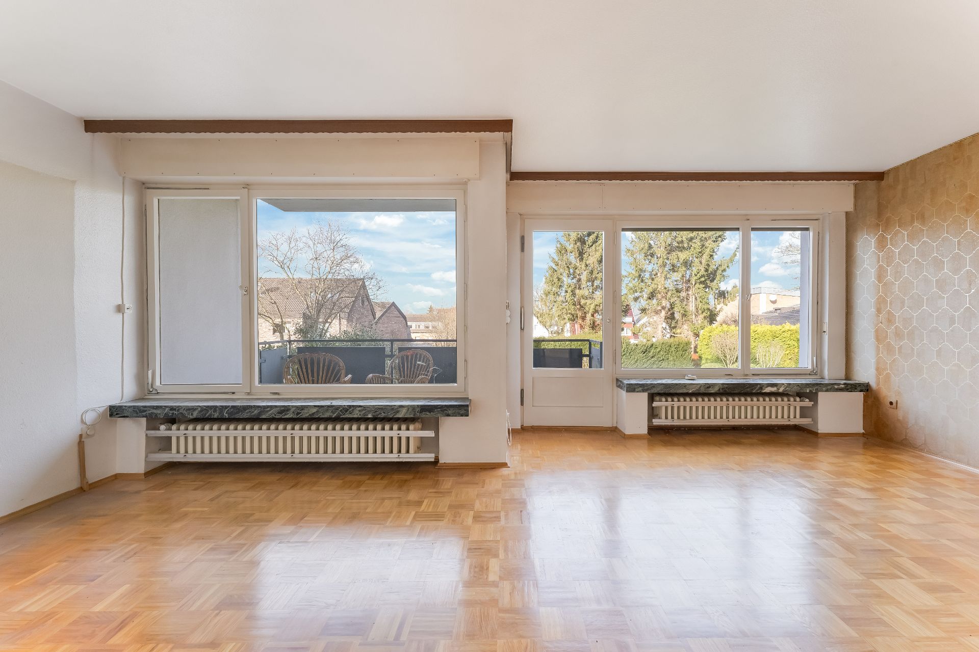 Helles Wohnzimmer mit Parkettboden, großen Fenstern und Blick auf Garten und Nachbarhäuser.