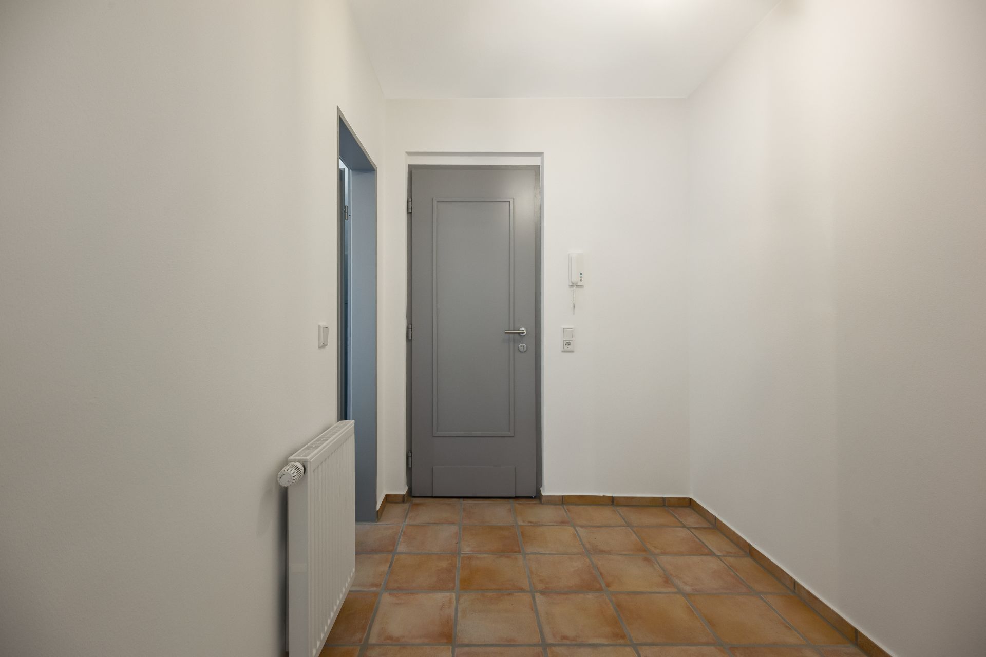 Heller Flur mit grauer Tür, weißer Wand, Fliesenboden und Heizkörper.