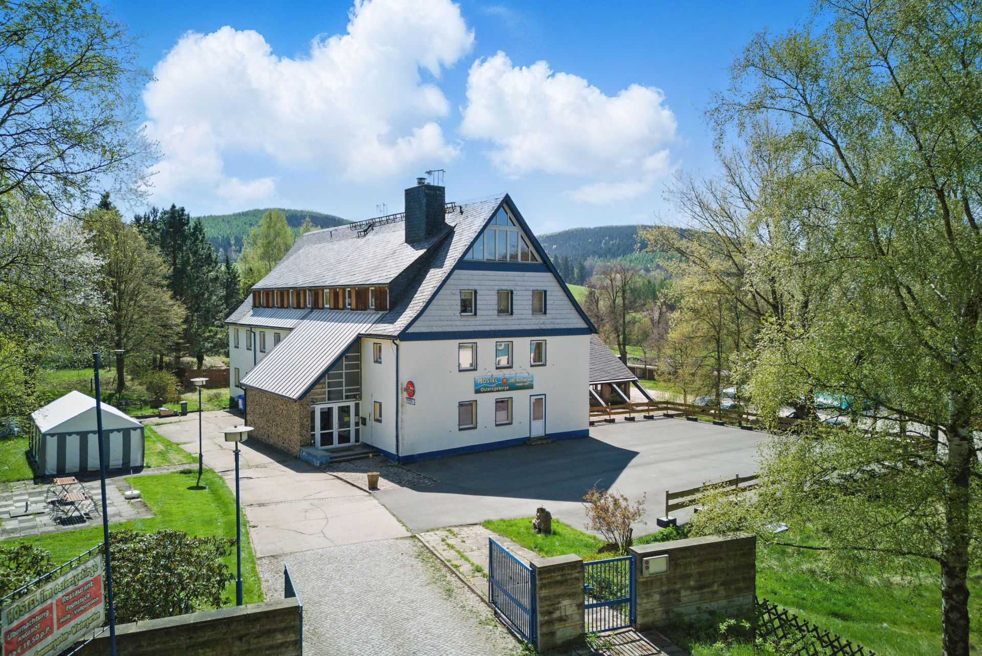 Großes weißes Wohnhaus mit dunklem Dach, umgeben von Bäumen und Hof, bei sonnigem Wetter.