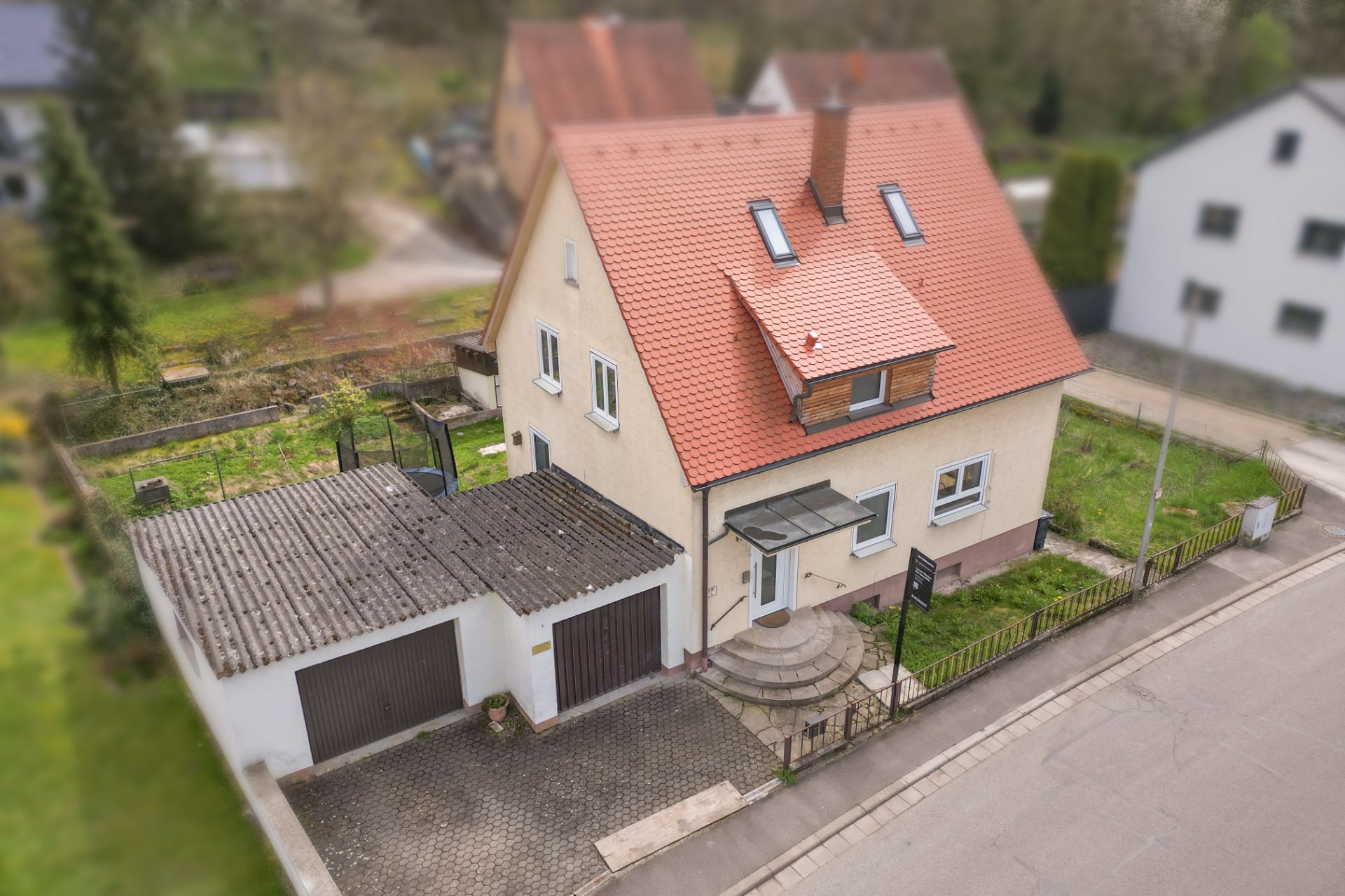 Zweistöckiges Haus mit rotem Dach, Garage und kleinem Garten an einer Straße.