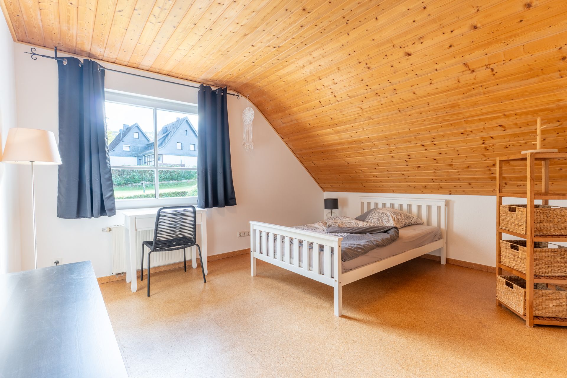 Helles Schlafzimmer mit Holzbett, Schreibtisch, Stuhl und Holzdecke im Dachgeschoss.
