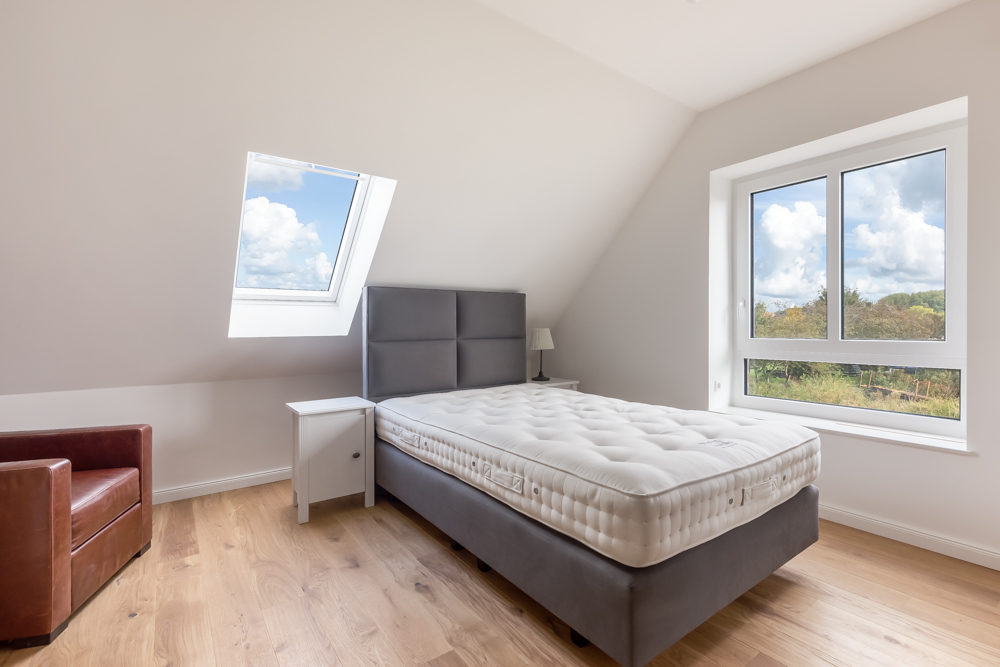 Helles Schlafzimmer mit Bett, Nachttisch, Lampe, Sessel und zwei Fenstern.