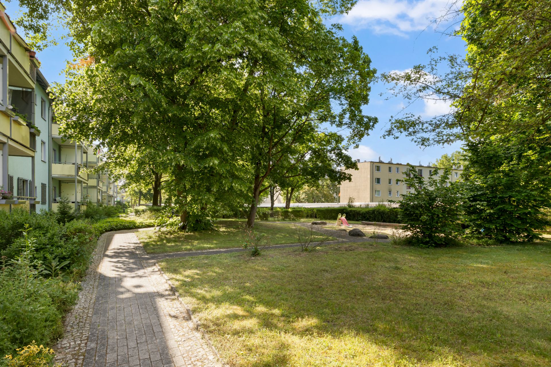 Gepflegter Garten mit Bäumen, Rasen und schmalem gepflastertem Weg neben Wohnhäusern.