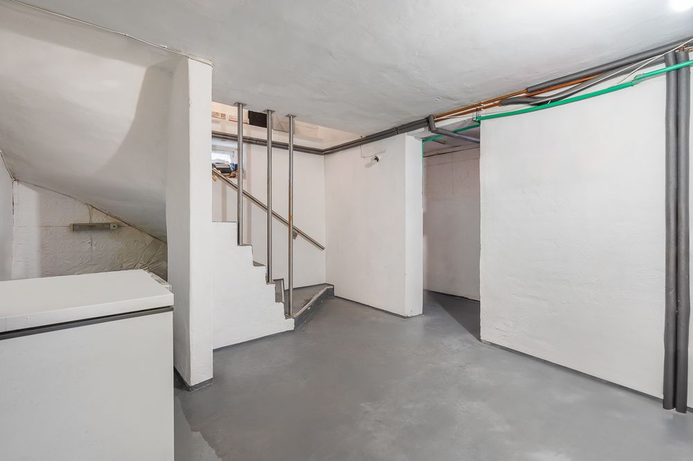 Kellerraum mit weißen Wänden, grauem Boden, Treppe und sichtbaren Rohren an der Decke.