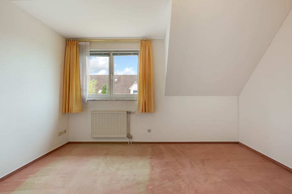 Leeres Zimmer mit schräger Decke, großem Fenster und gelben Vorhängen.