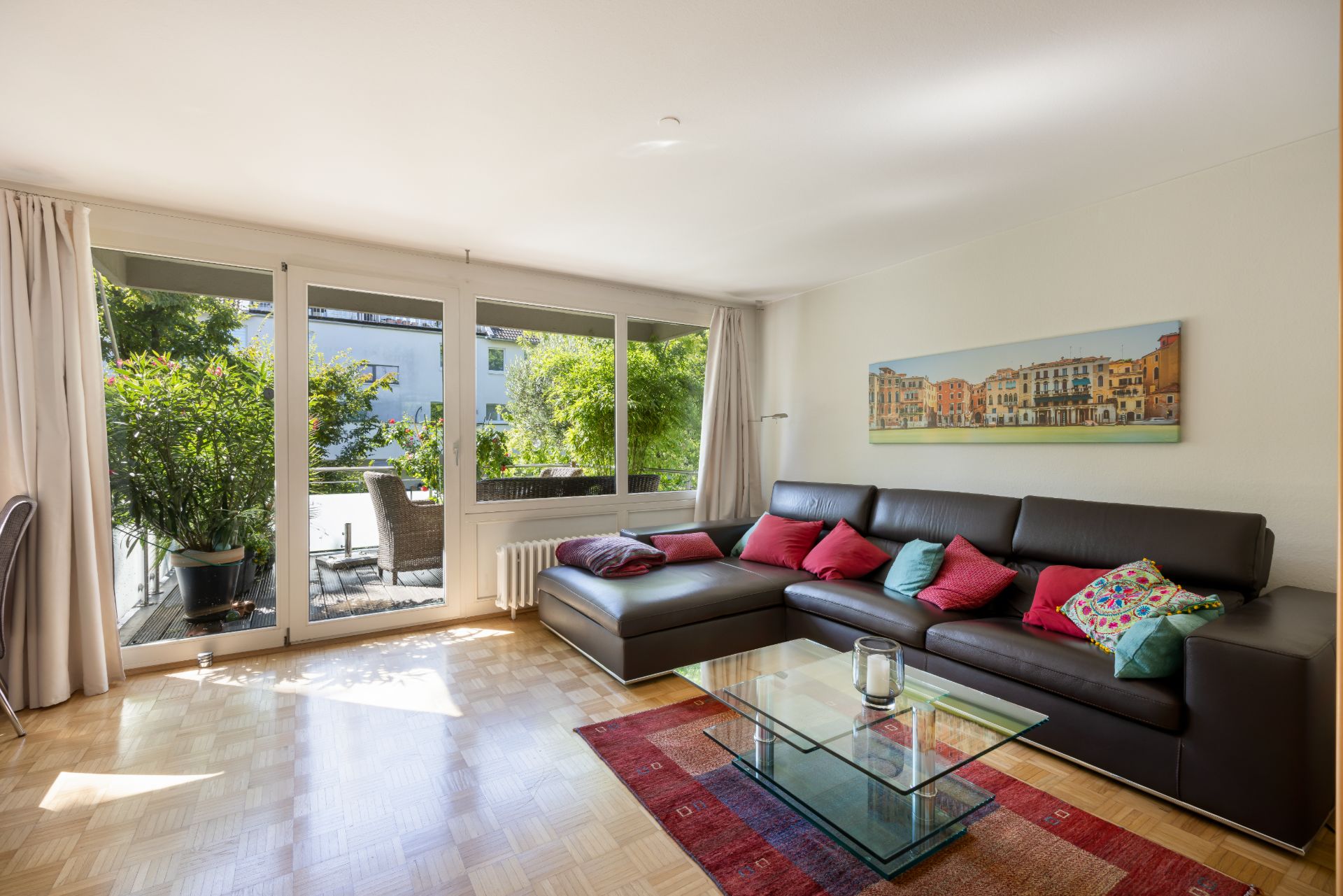 Helles Wohnzimmer mit Ecksofa, Glastisch, Teppich und Zugang zur Terrasse mit Gartenmöbeln.