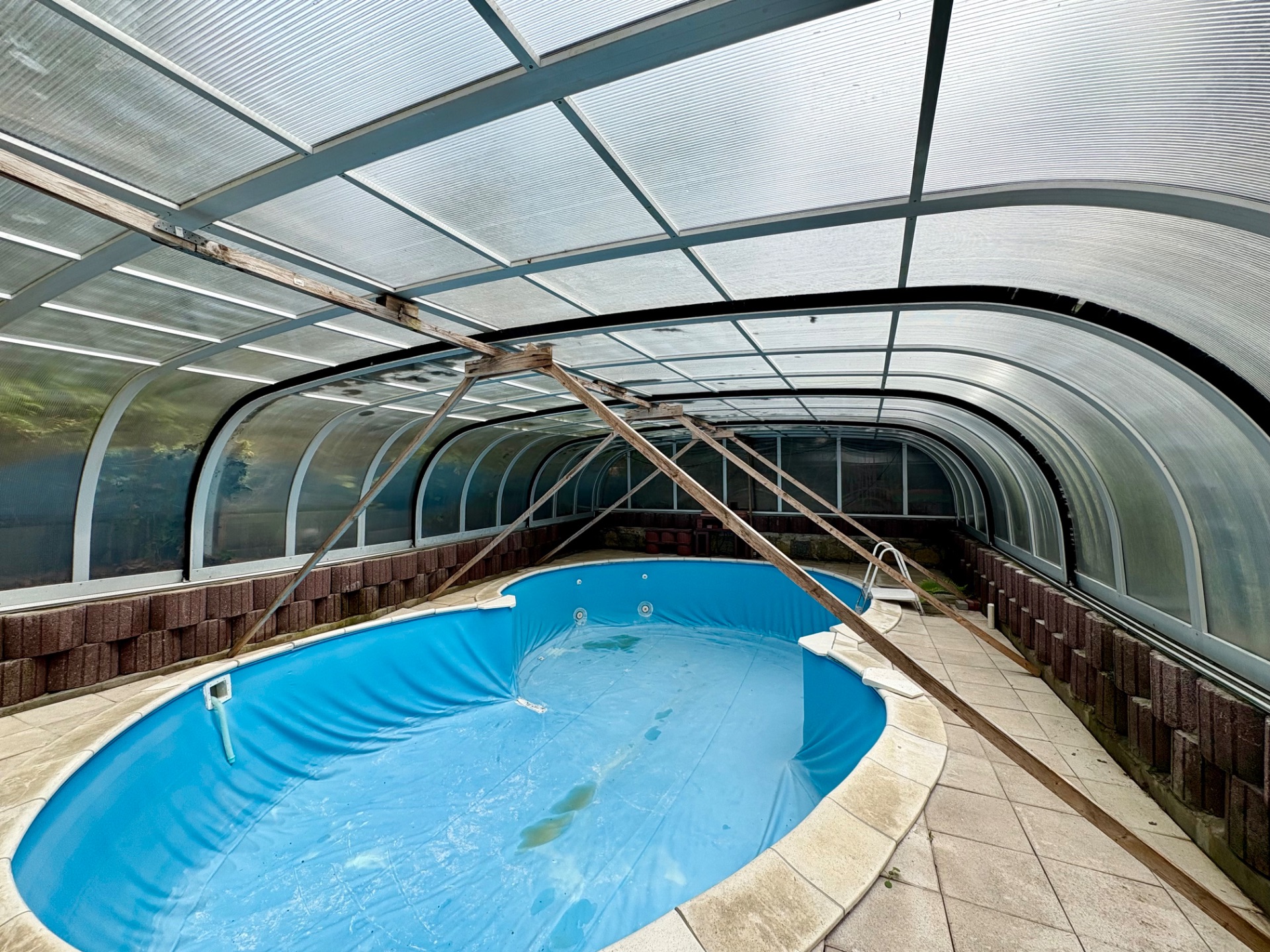 Überdachtes, leeres Schwimmbecken mit Fliesenboden und Glaswänden, mit Holzstreben gestützt.