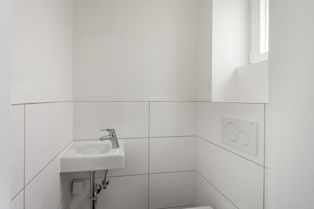 Kleines, weiß gefliestes WC mit kleinem Waschbecken und Fenster.