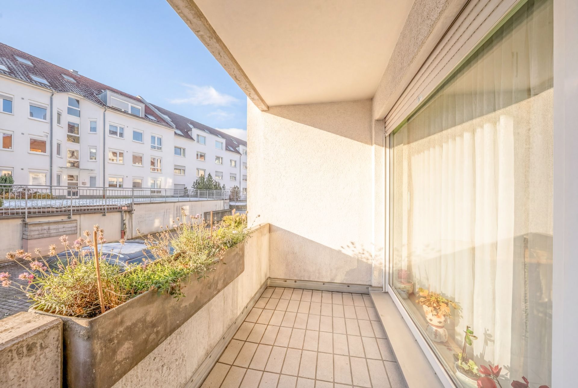 Heller Balkon mit Fliesenboden, Blumenkästen und Blick auf benachbarte Wohnhäuser.