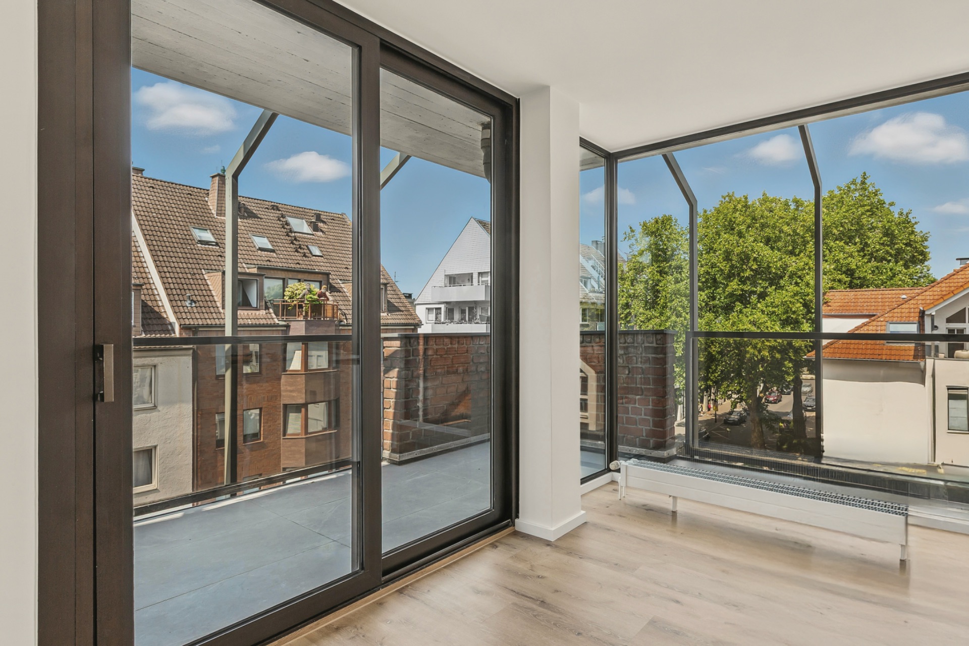 Heller Raum mit großen Fenstern und Zugang zu einem Balkon mit Blick auf Nachbarhäuser und Bäume.