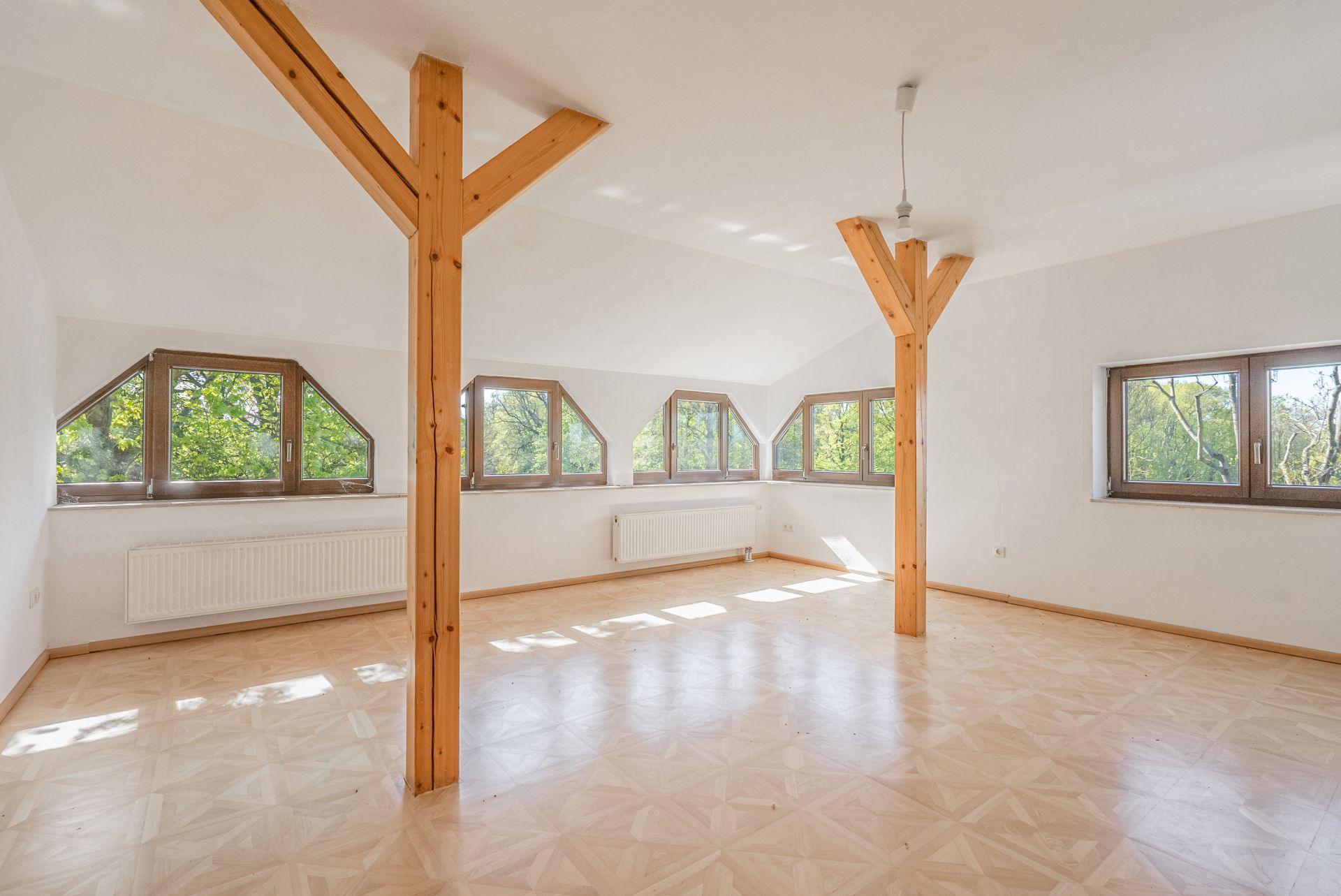 Heller, leerer Raum mit Holzstützen, großen Fenstern und Parkettboden.