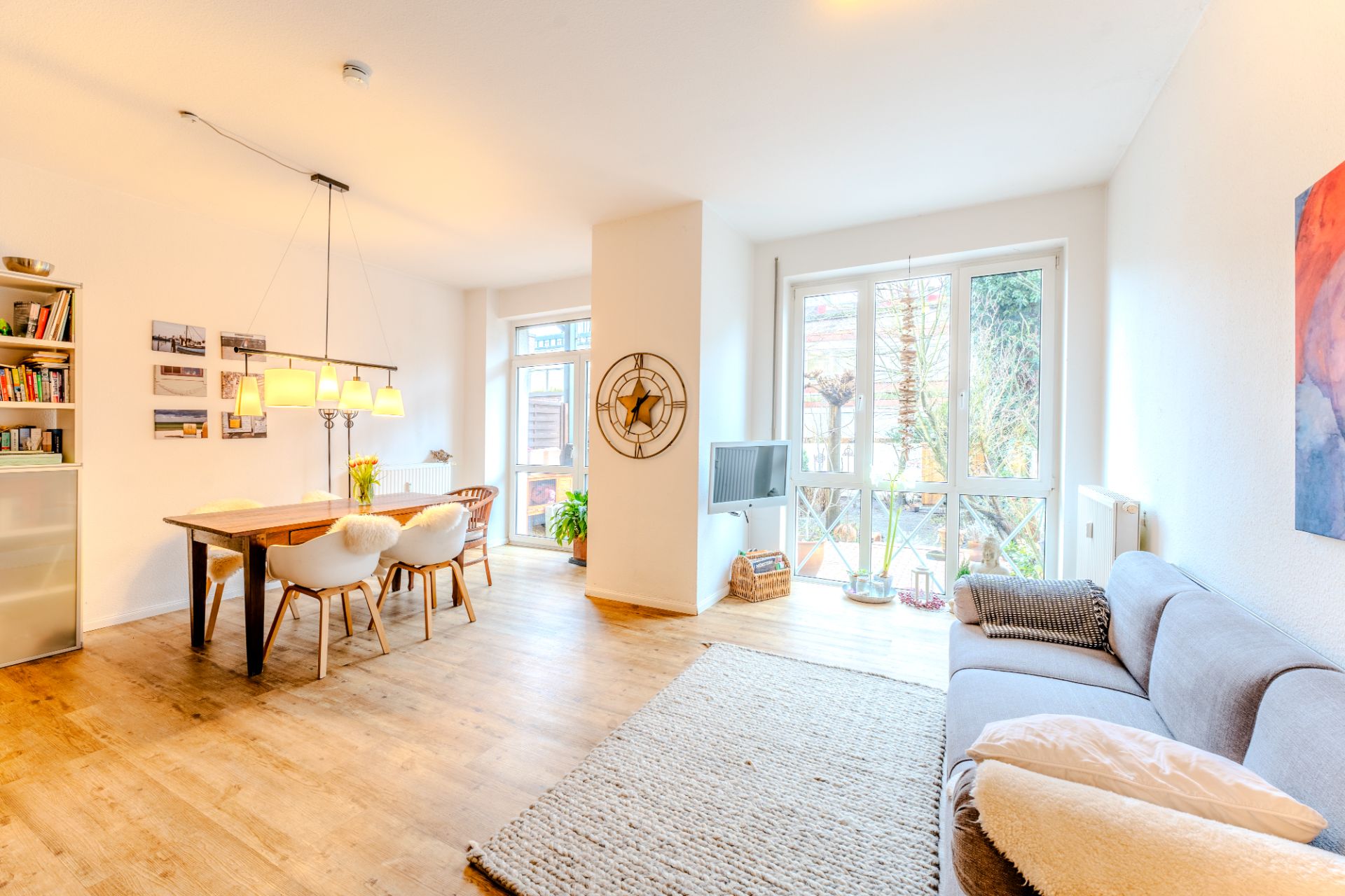 Helles Wohnzimmer mit grauem Sofa, Esstisch, großer Fensterfront und Holzboden.