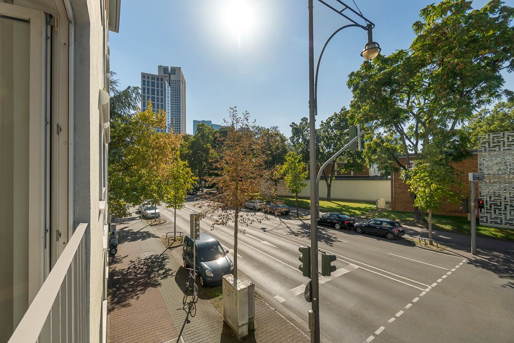 Blick vom Balkon auf eine sonnige Straße mit Bäumen, Autos und Gehweg in der Stadt.