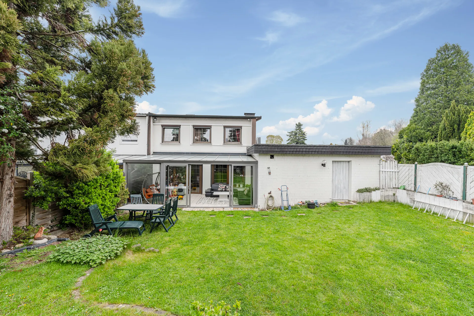 Grüner Garten mit Terrasse und Sitzgruppe, Blick auf ein weißes Wohnhaus mit großen Fenstern.