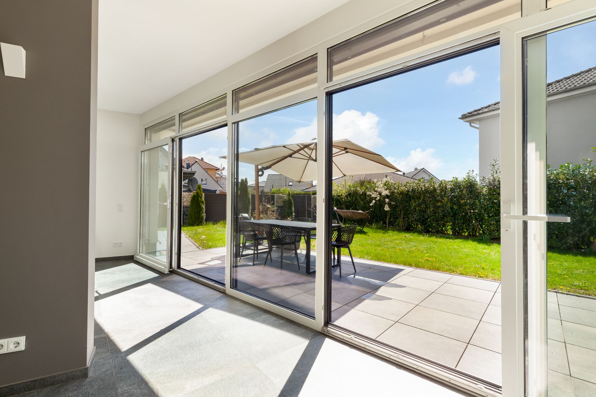 Helle Terrasse mit Glasfront, Garten, Tisch, Stühlen und großem Sonnenschirm.
