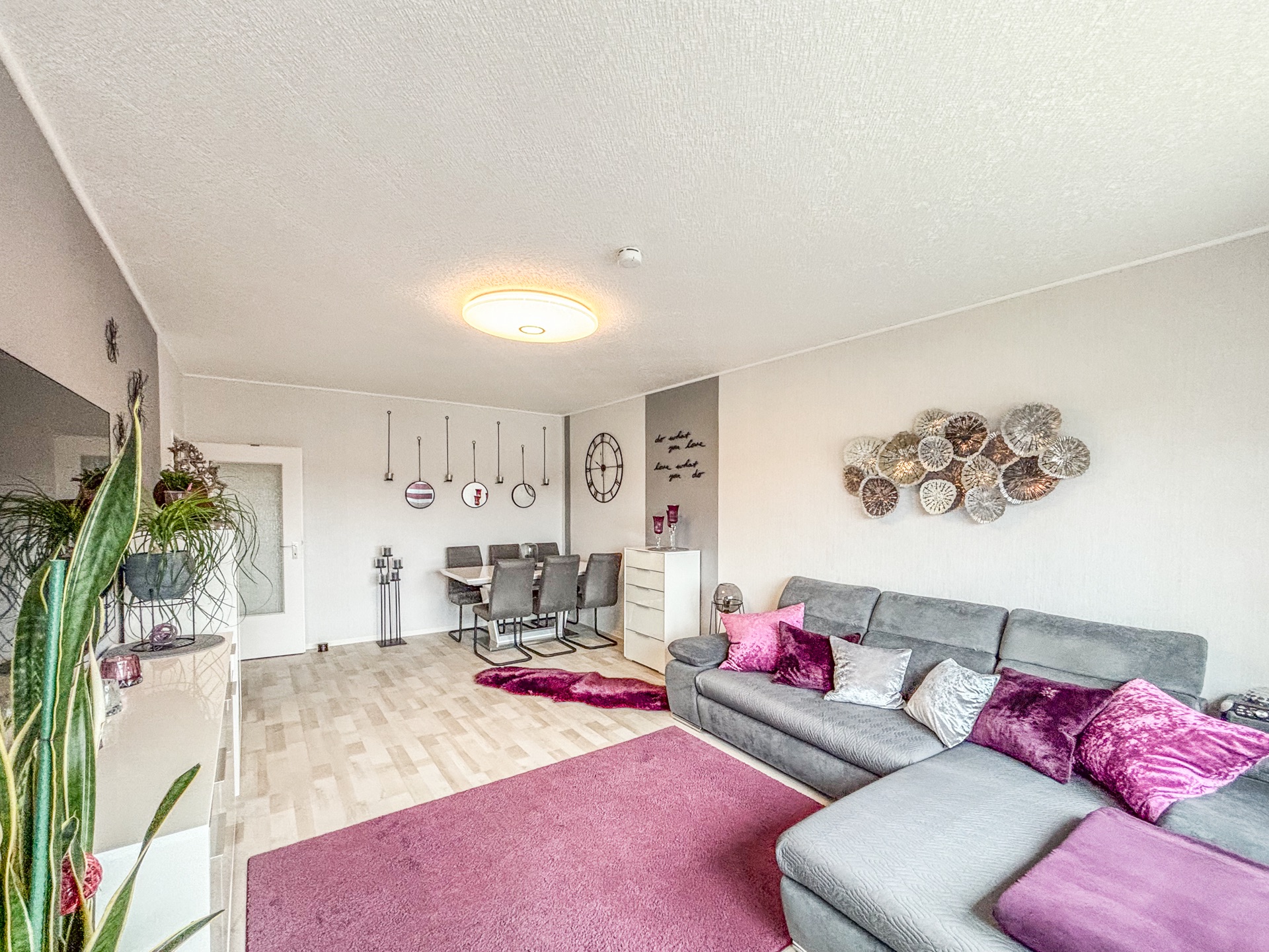 Modernes Wohnzimmer mit grauem Sofa, violetten Kissen, Esstisch und dekorativer Wandkunst.
