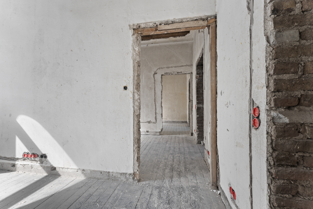 Leerstehender Raum mit weißen Wänden, Holzboden und sichtbaren Steckdosen in Renovierung.