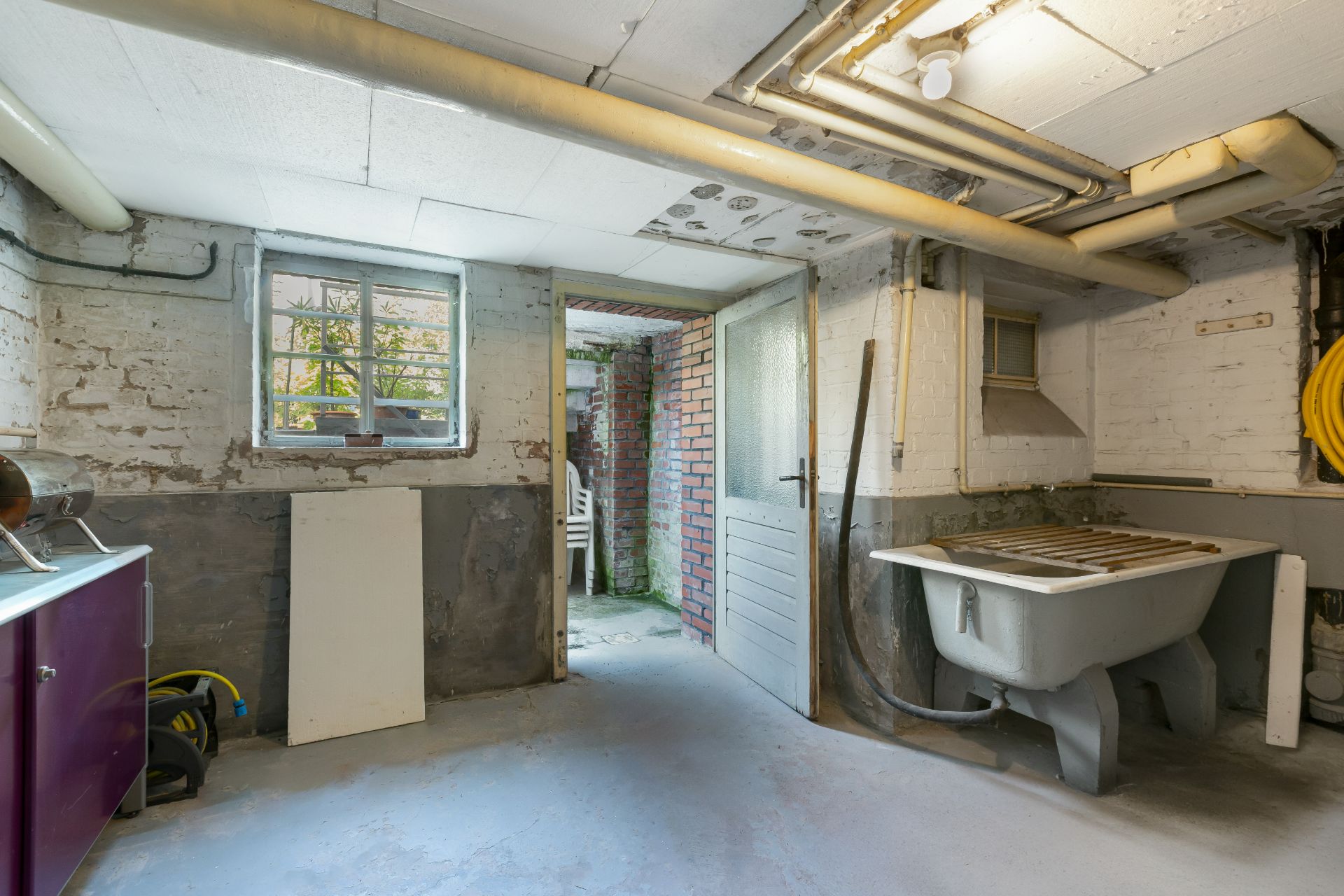 Kellerraum mit Waschbecken, offenem Fenster und sichtbaren Rohren an der Decke.