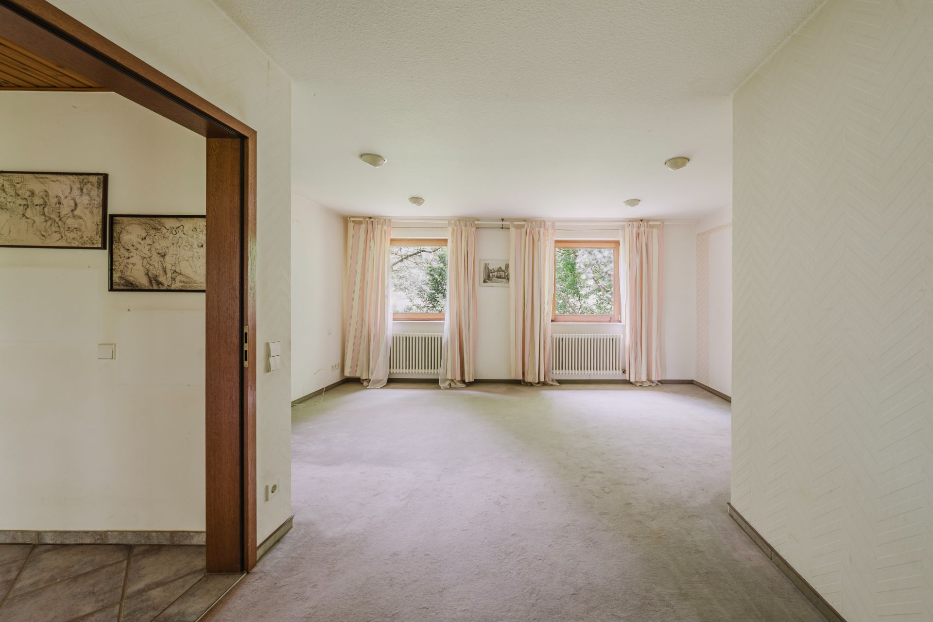 Leeres Wohnzimmer mit zwei Fenstern, hellen Vorhängen und Teppichboden.