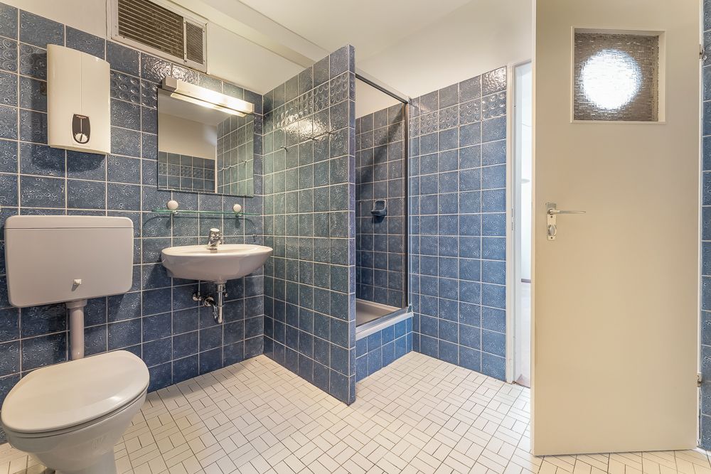 Badezimmer mit blauen Fliesen, Toilette, Waschbecken, Spiegel und Dusche.