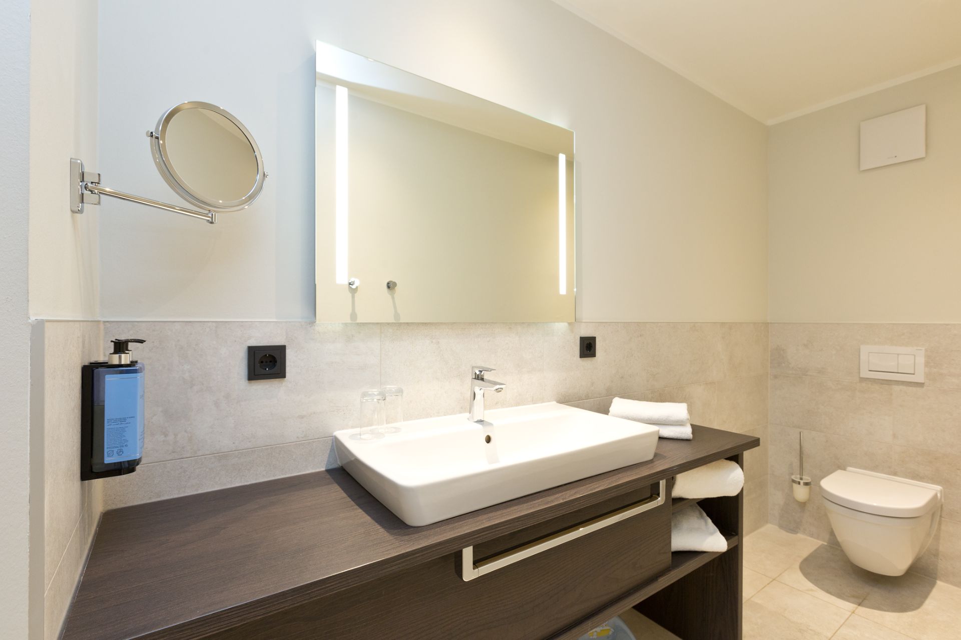 Modernes Badezimmer mit Waschbecken, beleuchtetem Spiegel, Handtüchern und wandhängender Toilette.
