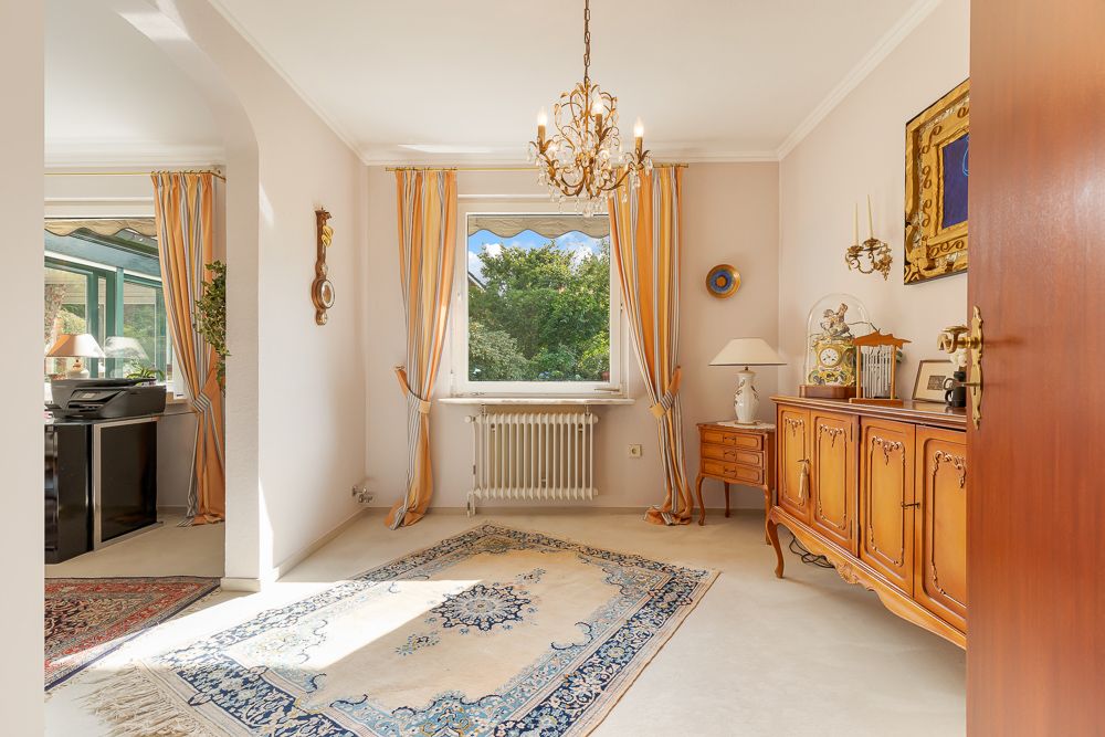 Helles Wohnzimmer mit Teppich, Kronleuchter und Holzkommode vor einem Fenster mit Vorhängen.