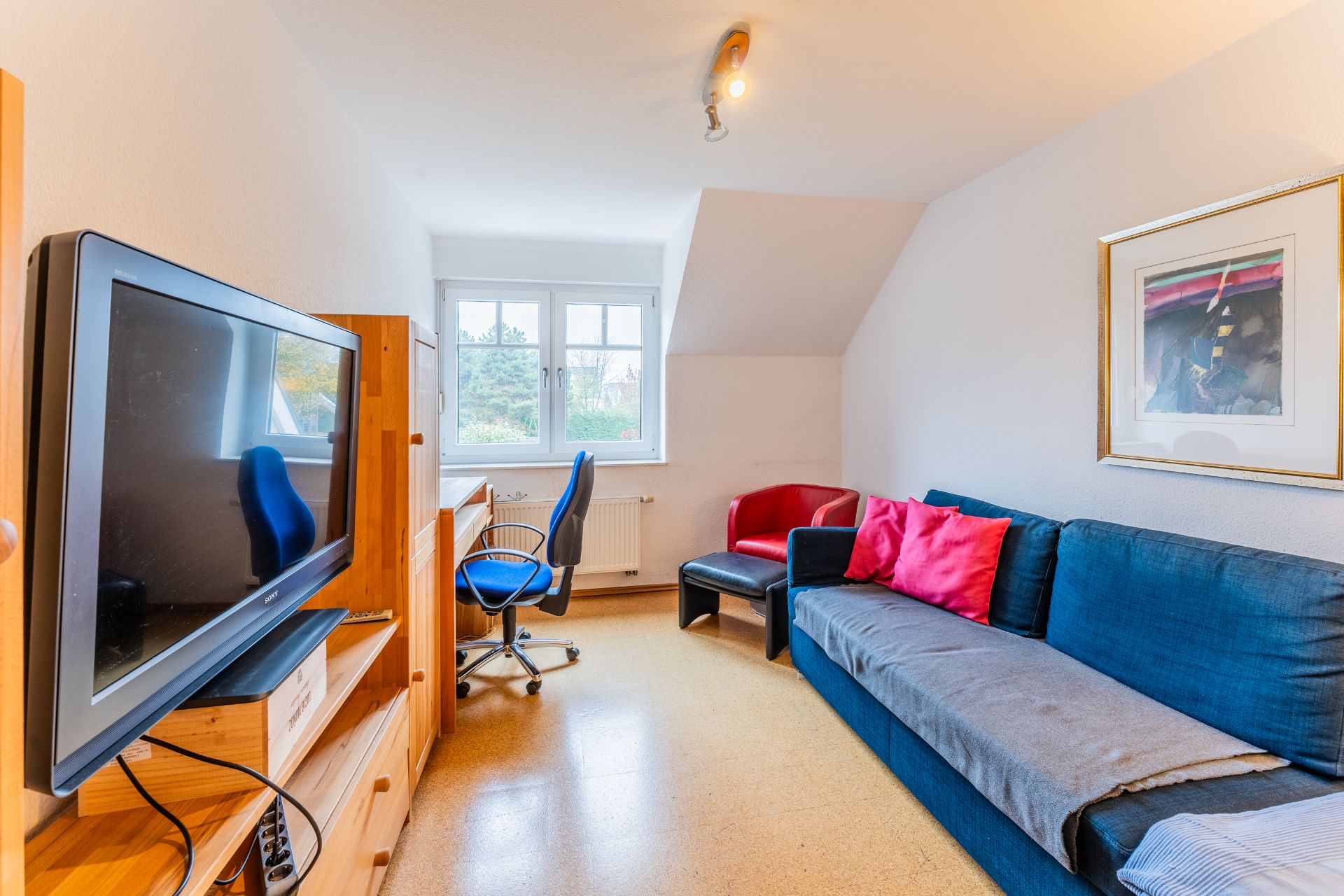 Helles Wohnzimmer mit blauem Sofa, rotem Sessel, Fernseher, Schreibtisch und Fenster.