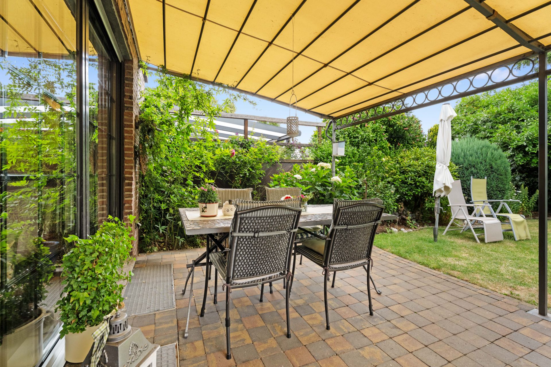 Überdachte Terrasse mit Tisch und Stühlen, umgeben von viel Grün und Gartendekoration.