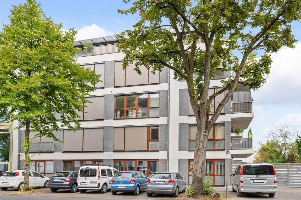 Mehrstöckiges Wohnhaus mit großen Fenstern, Bäumen und parkenden Autos davor.