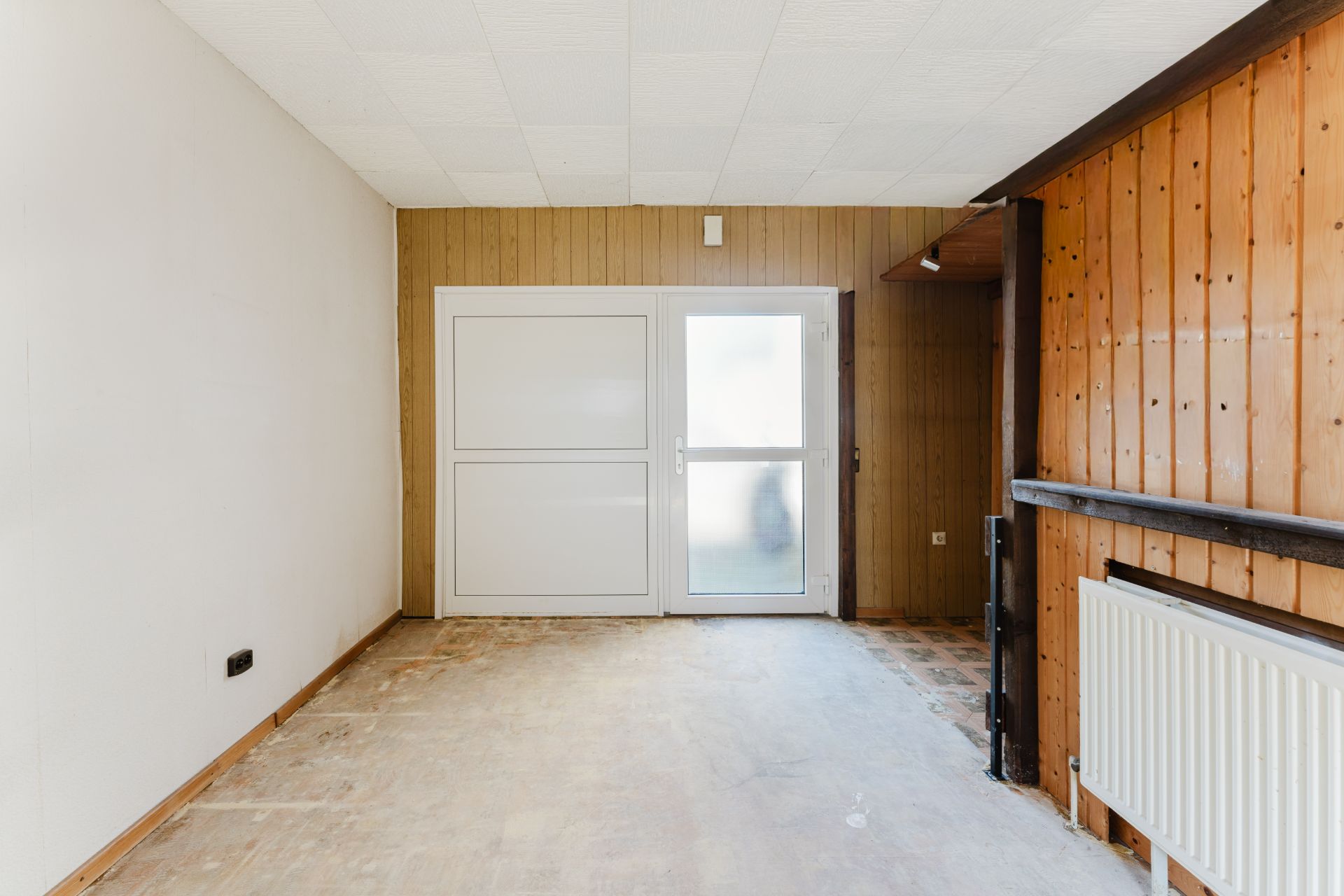 Leerer Raum mit Holzwänden, Fenster und Tür, Heizkörper an der rechten Wand.