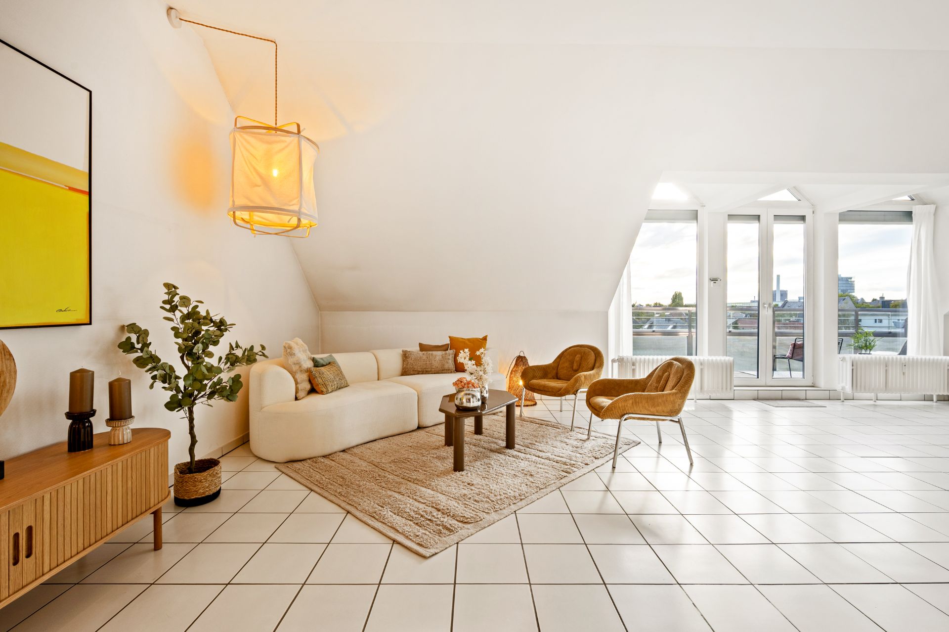 Helles Wohnzimmer mit weißem Sofa, Teppich, Holztisch, zwei Stühlen und großen Fenstern mit Blick nach draußen.
