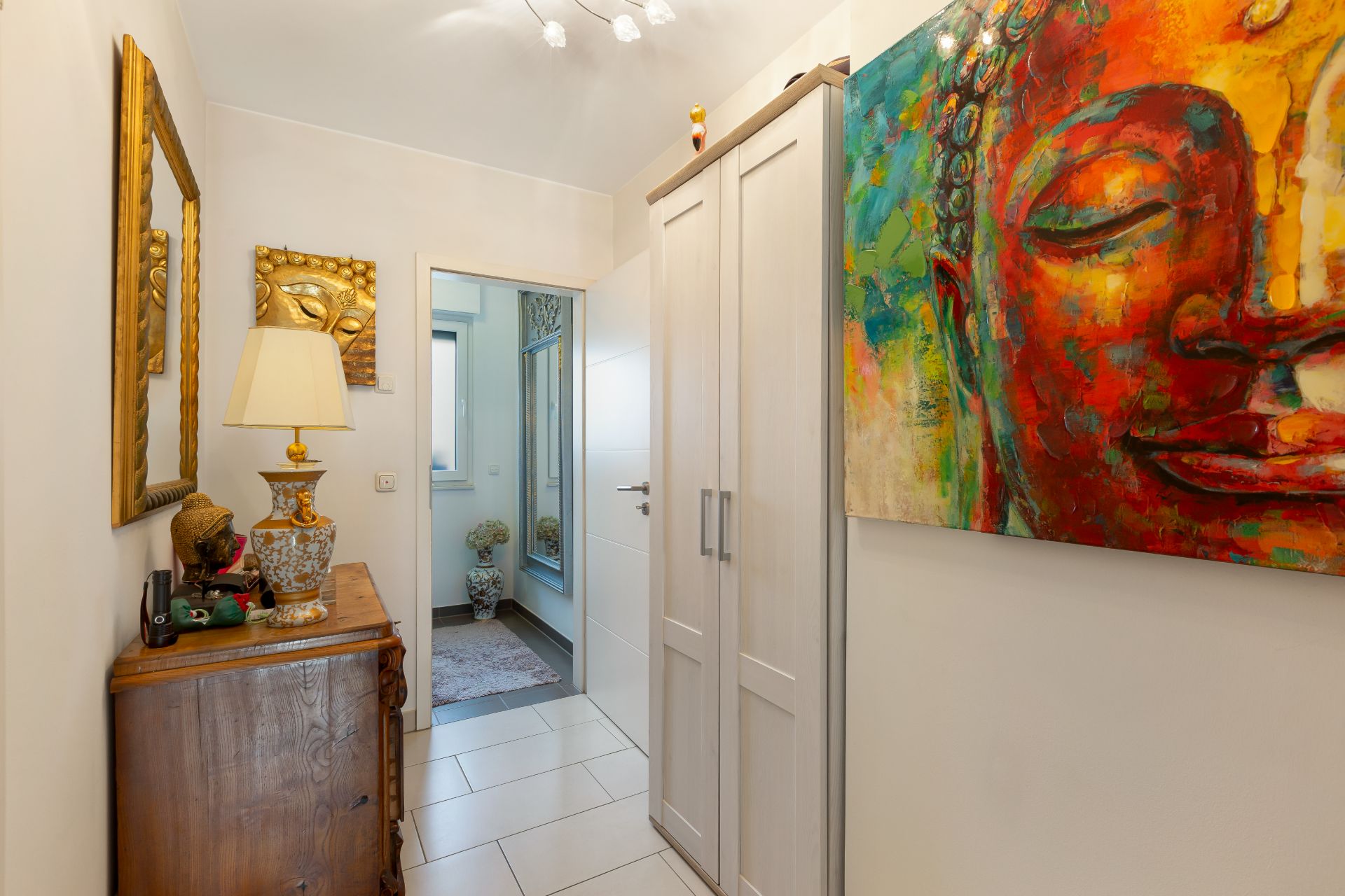 Flur mit buntem Buddha-Gemälde, Holzkommode, Spiegel und weißem Schrank.