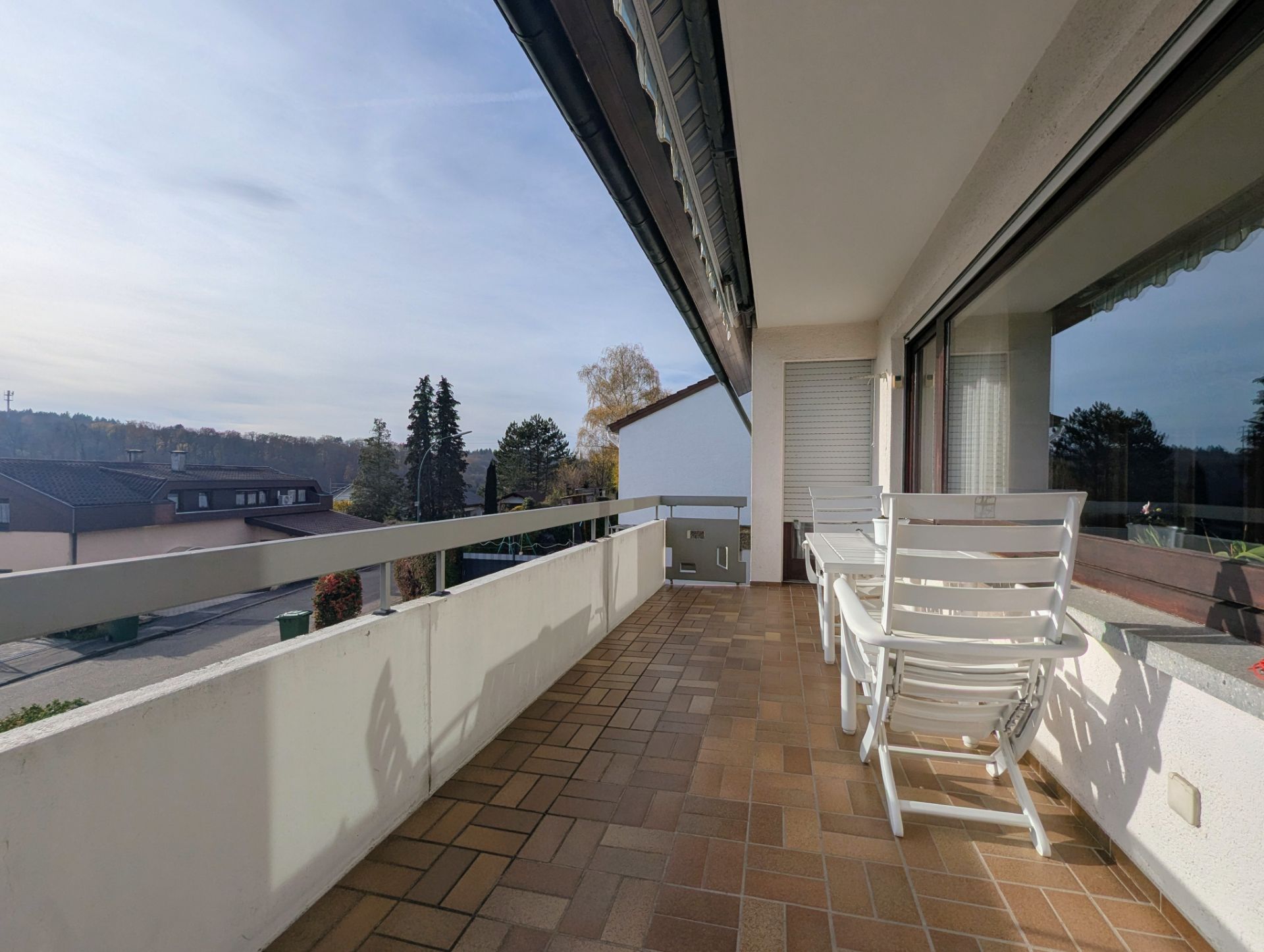 Balkon mit zwei weißen Stühlen und einem Tisch, Blick auf Wohnhäuser und Bäume.