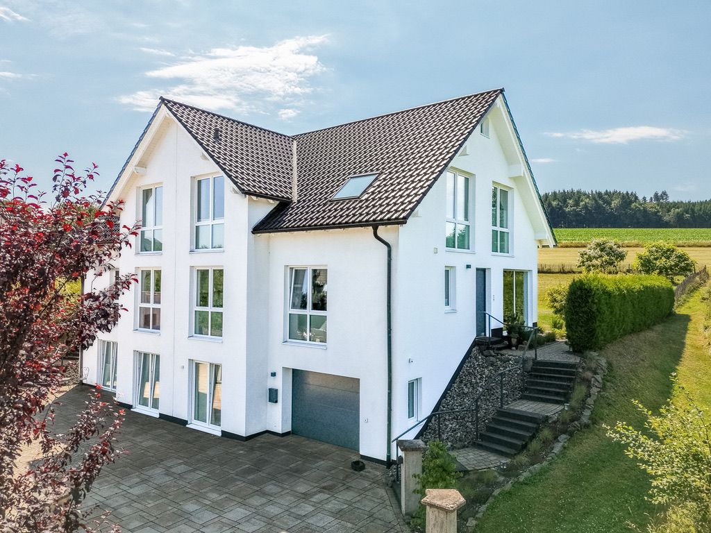 Modernes weißes Einfamilienhaus mit großen Fenstern und dunklem Satteldach auf dem Land.