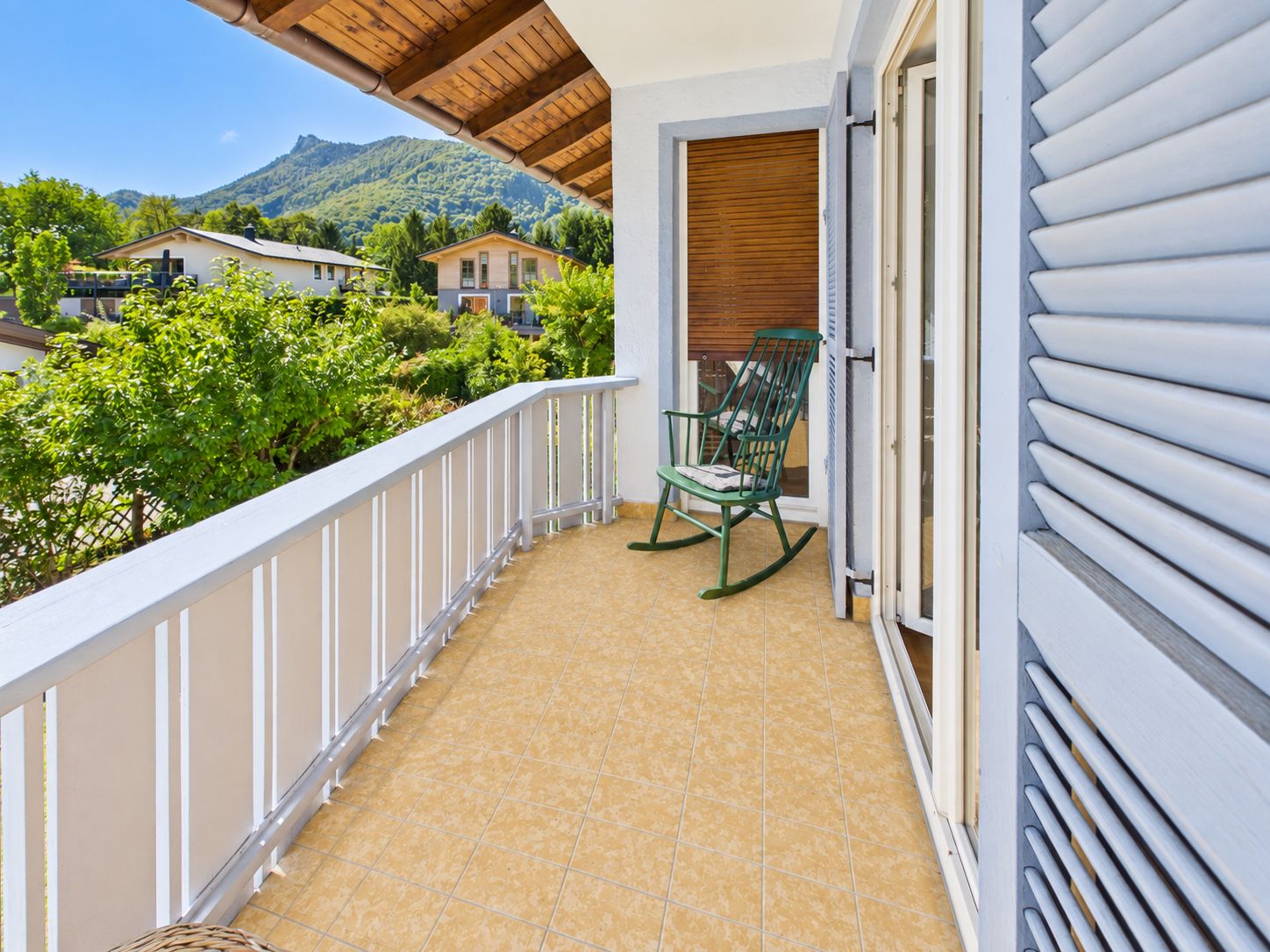 Überdachter Balkon mit Schaukelstuhl, Blick auf Bäume und Nachbarhäuser, Berge im Hintergrund.