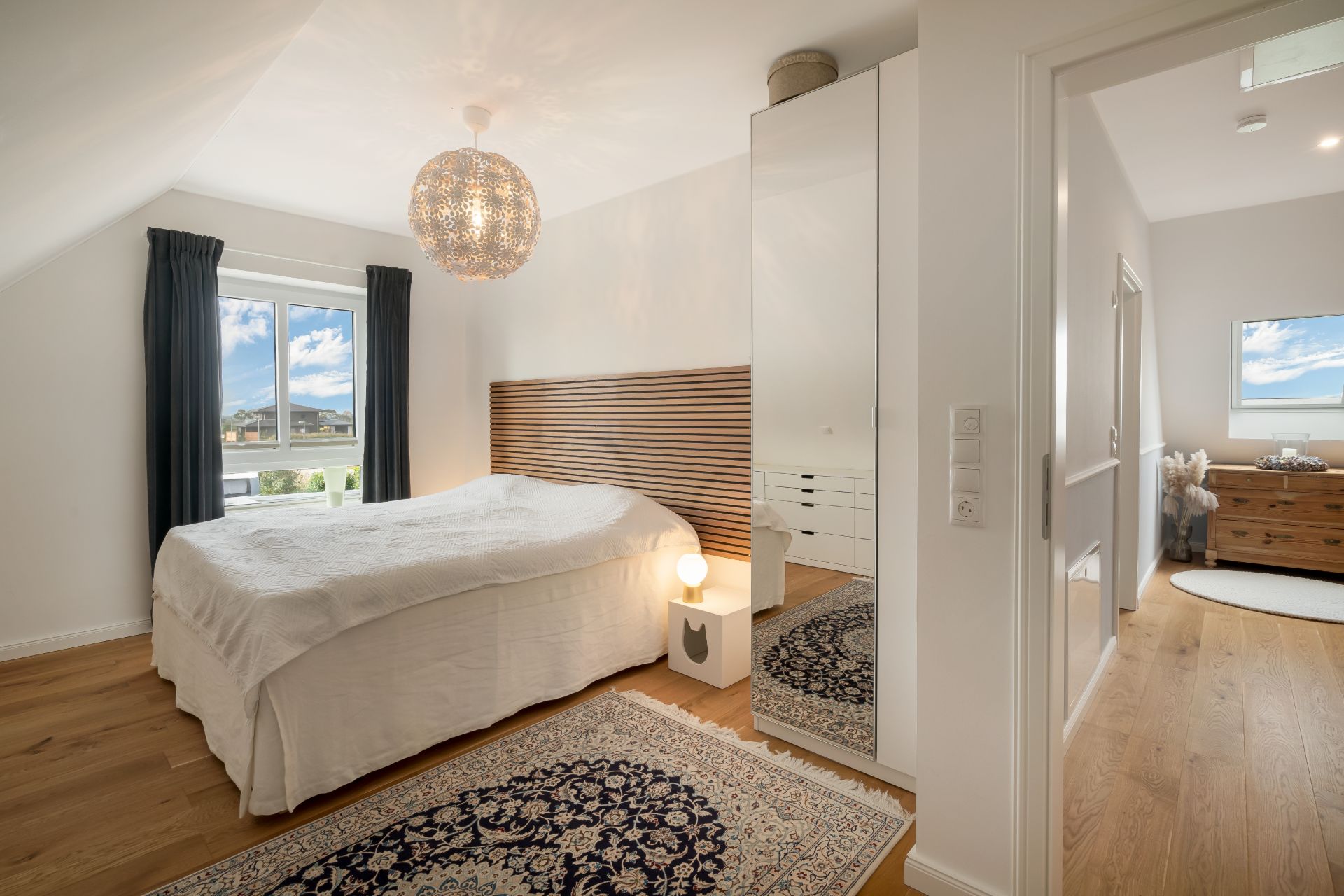 Helles Schlafzimmer mit Doppelbett, Holzfußboden, Teppich, Spiegelschrank und Fenster mit Aussicht.