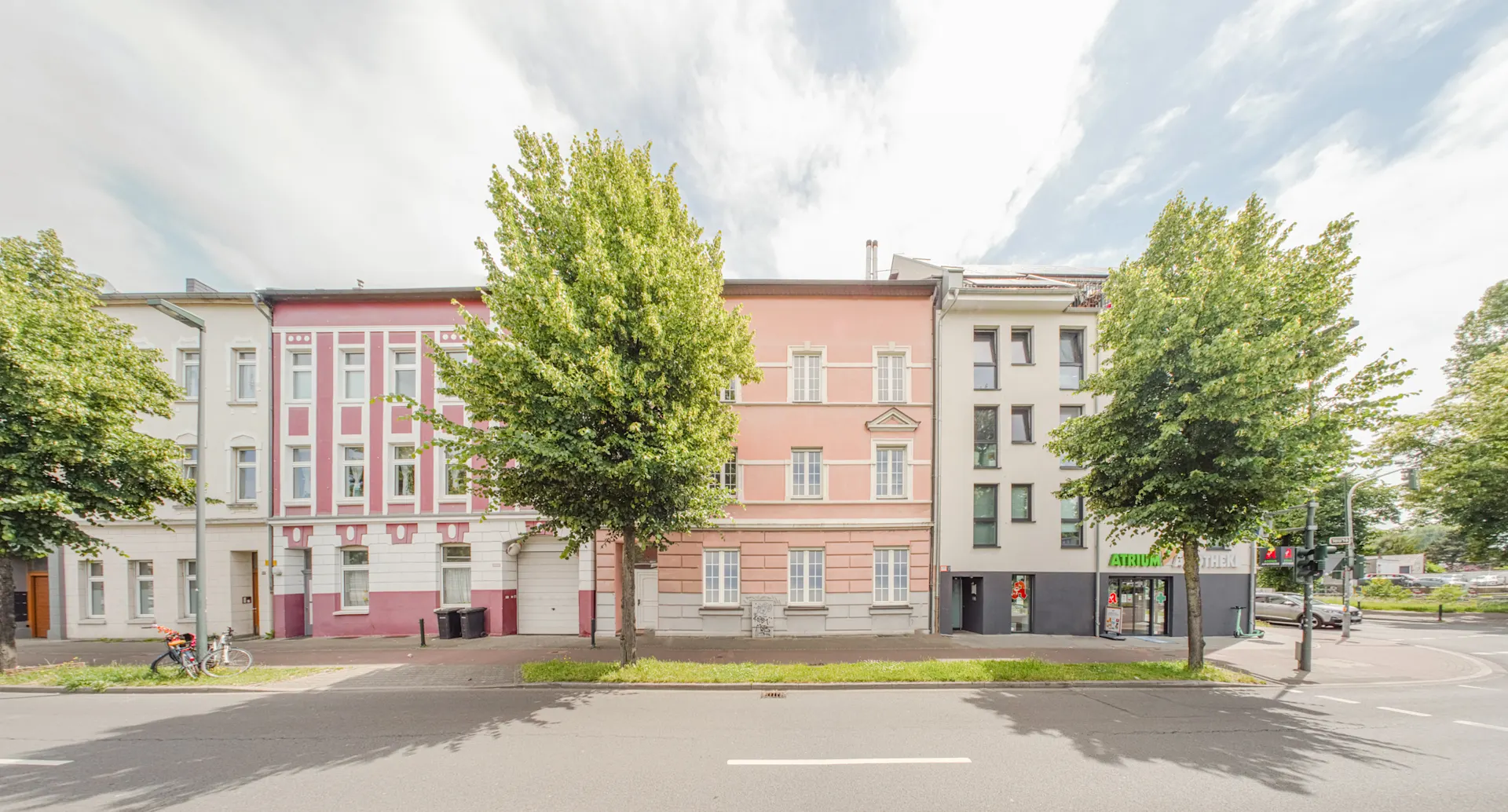 Mehrere Wohnhäuser mit Bäumen davor an einer Straße, darunter ein rosafarbenes Gebäude.