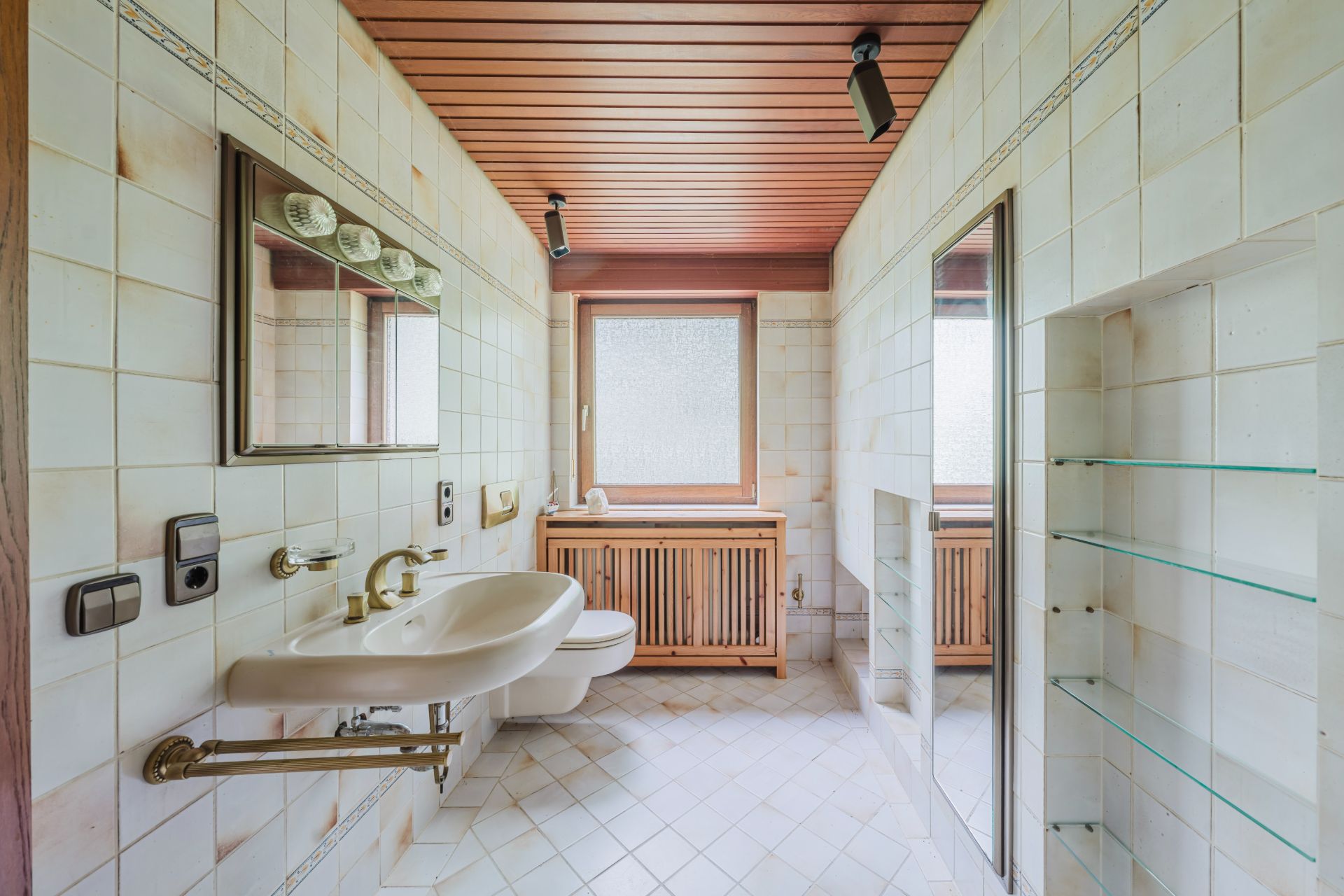 Helles Badezimmer mit Waschbecken, Spiegel, WC, Fenster und Glasregalen an der Wand.