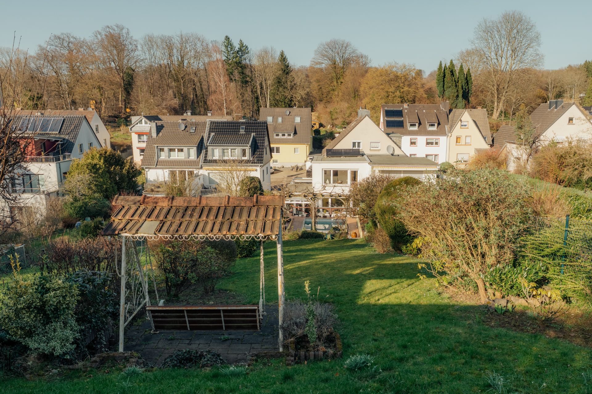Blick auf einen Garten mit Holzpergola und Sitzbank, im Hintergrund mehrere Wohnhäuser.