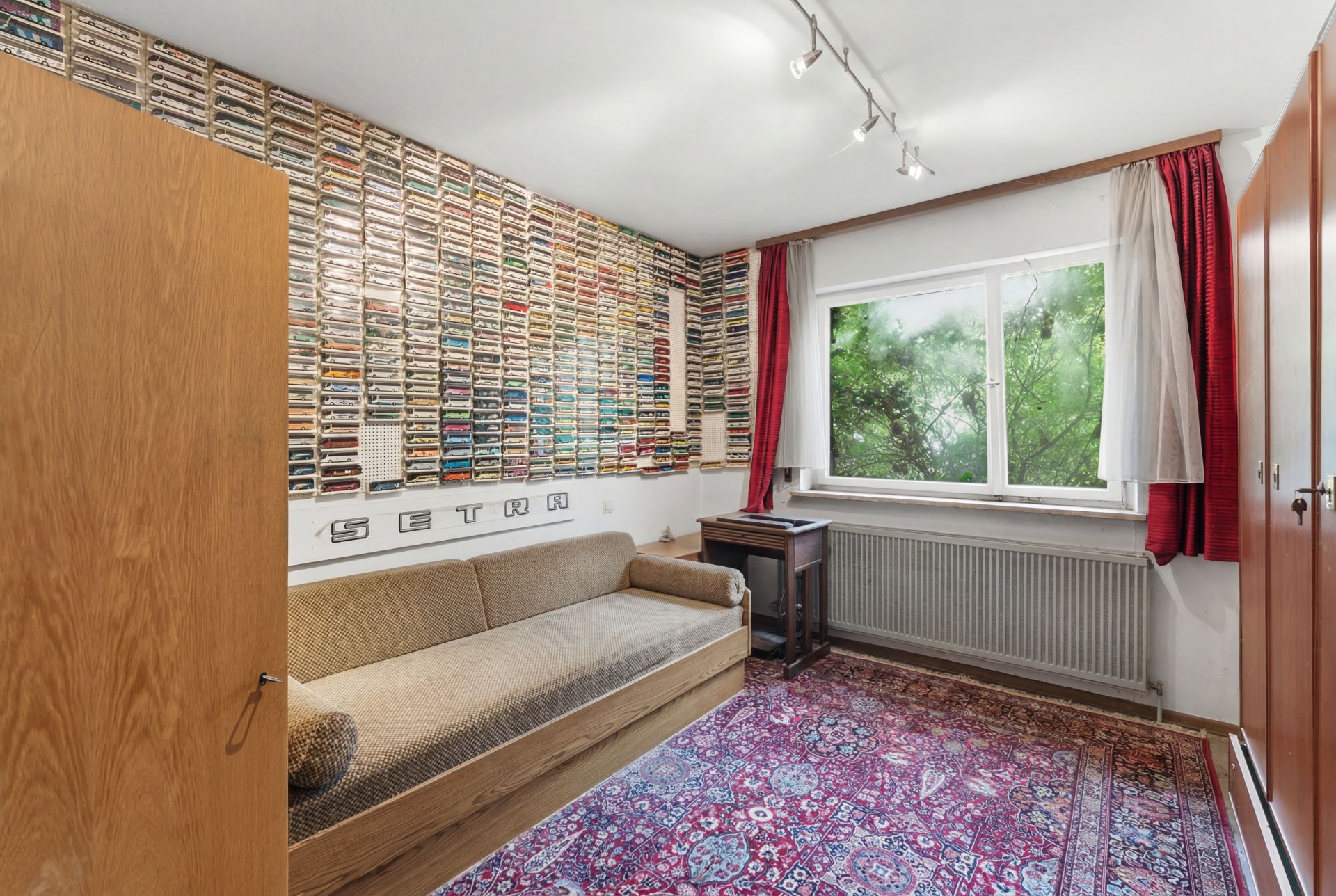 Zimmer mit Sofa, buntem Teppich, Regalwand voller Kassetten und großem Fenster mit roten Vorhängen.