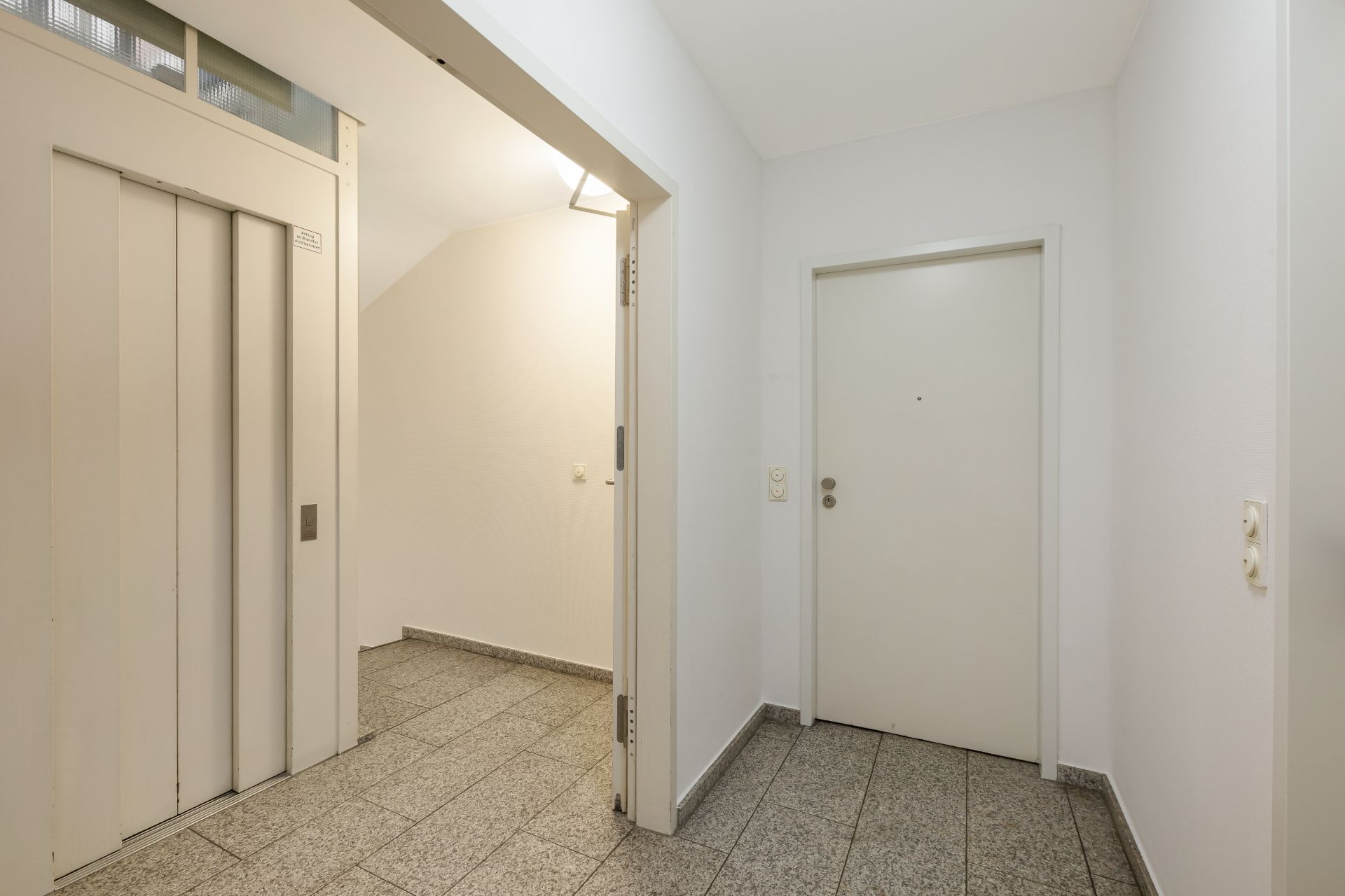 Hausflur mit Aufzug und weißer Eingangstür, gefliestem Boden und weißen Wänden.