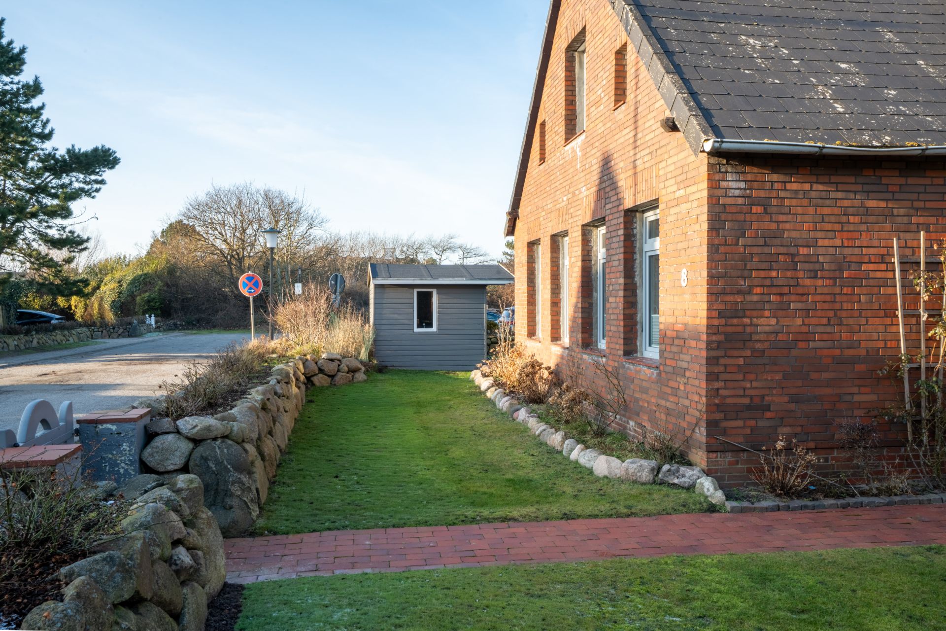 Backsteinwohnhaus mit Garten, Rasen und gepflastertem Weg, daneben ein kleines graues Gartenhaus.