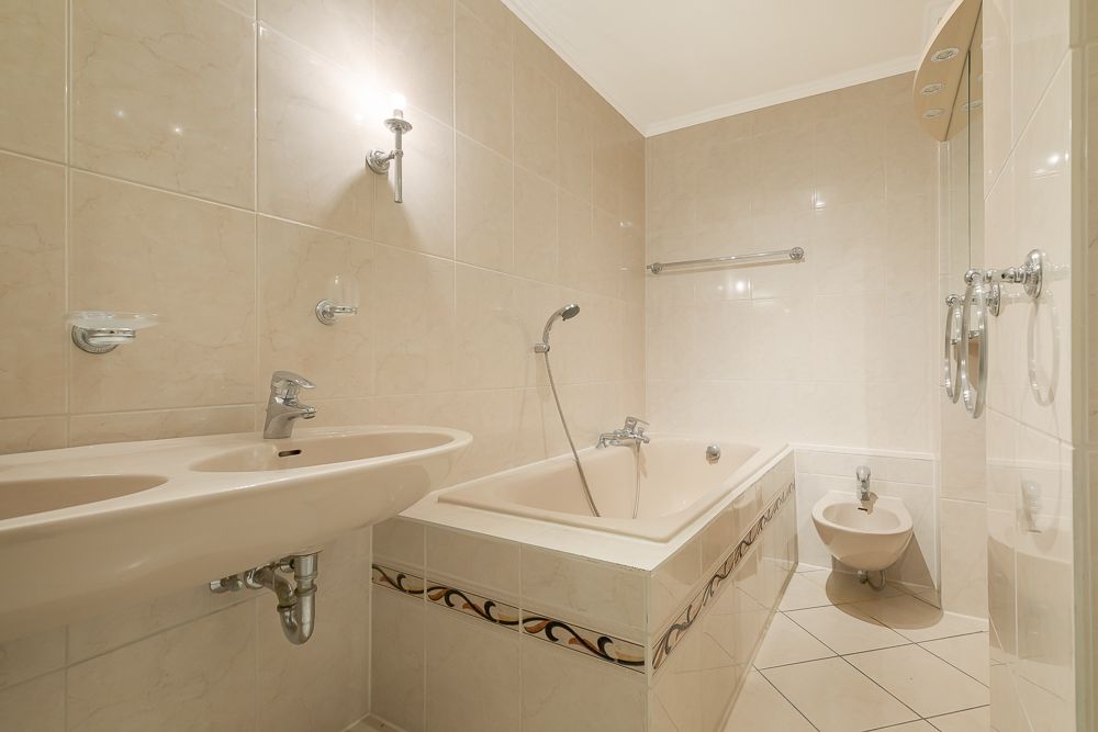 Helles Badezimmer mit Badewanne, Doppelwaschbecken und Bidet, alles mit beigen Fliesen.