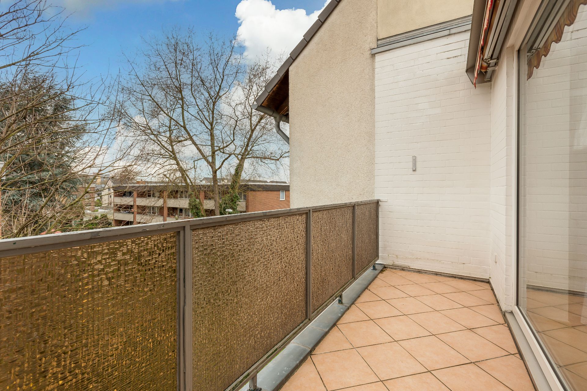 Balkon mit braunem Geländer und Fliesenboden, Ausblick auf Bäume und andere Gebäude.