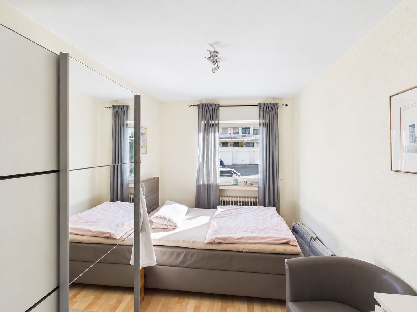Helles Schlafzimmer mit Doppelbett, Spiegelschrank und zwei Fenstern mit grauen Vorhängen.