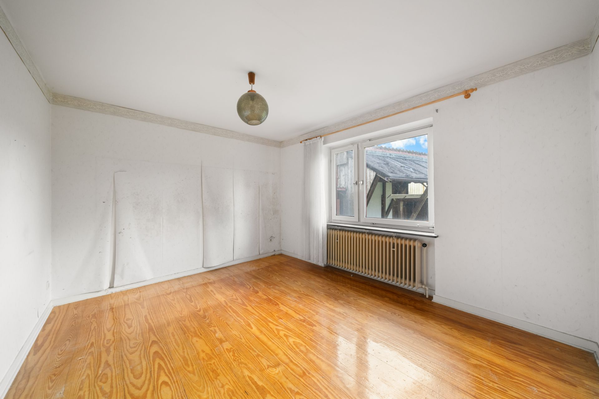 Leerer Raum mit Holzboden, weißem Wandanstrich, großem Fenster und Heizung unter dem Fenster.